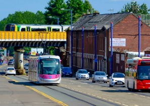 Image shows cars, buses, the tram on a road, with a train running across a bridge behind them
